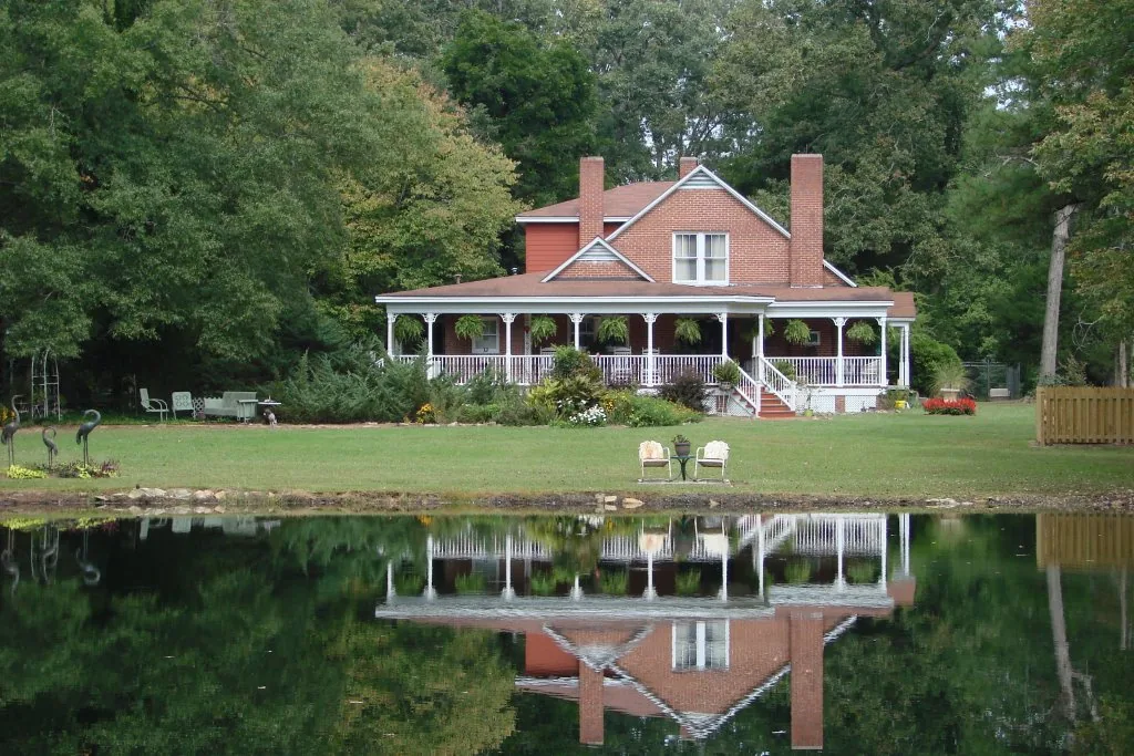 The brick Cape Cod house with its white wrap-around porch, reflected in the pond at Heartfriends Inn